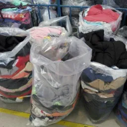 Przezroczyste worki wypełnione różnorodną odzieżą używaną, widoczne warstwy ubrań w różnych kolorach i wzorach, ustawione na betonowej podłodze w magazynie.