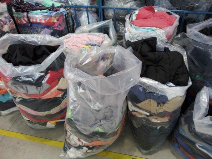 Przezroczyste worki wypełnione różnorodną odzieżą używaną, widoczne warstwy ubrań w różnych kolorach i wzorach, ustawione na betonowej podłodze w magazynie.