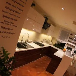 Nowoczesna kuchnia z ciemnobrązowymi szafkami dolnymi i białymi górnymi, widok na zlew, płytę grzewczą i ścianę z napisem motywacyjnym, sfotografowana z szerokiego kąta.