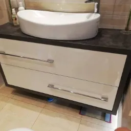 Łazienkowa szafka pod umywalkę z dwiema szufladami w kolorze białym, z ciemnoszarym blatem i chromowanymi uchwytami, stojąca na jasnej podłodze z płytek.