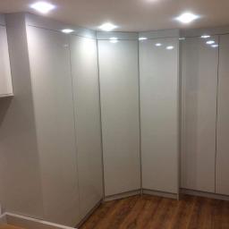 P.P.H Mag - Zabudowa narożna z białych, lakierowanych szaf z delikatnymi uchwytami, dopasowana do niskiego sufitu, z widoczną drewnopodobną podłogą.