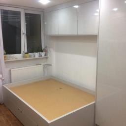 P.P.H Mag - Wnęka z szarą bazą łóżka z szufladami, jasnym drewnianym dnem, białe szafki wiszące i wysoka szafa w zabudowie, widok na okno z roślinami.