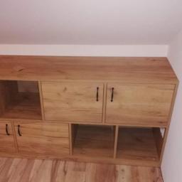 P.P.H Mag - Zabudowa meblowa pod skosem na poddaszu, z widocznymi półkami otwartymi i szafkami z ciemnymi uchwytami, na podłodze panele drewnopodobne, tuba silikonu budowlanego i butelka płynu w jednej z półek.