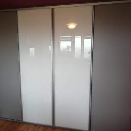 P.P.H Mag - Duża, czterodrzwiowa szafa przesuwna z szarymi i białymi panelami, widoczna na tle drewnianej podłogi i bordowej ściany, z odbiciem okna w białych panelach.