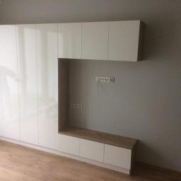 P.P.H Mag - Biała, lakierowana zabudowa ścienna z elementami drewnopodobnymi, obejmująca szafki i wnękę na sprzęt RTV, zamontowana na tle szarej ściany z widocznymi gniazdkami.