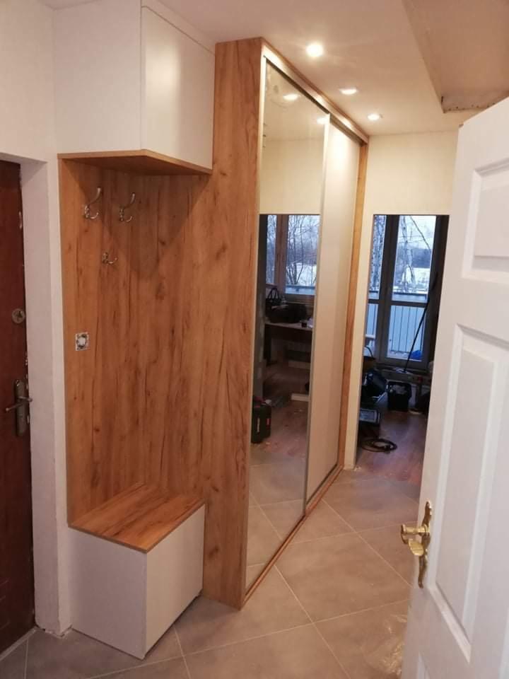 Przedpokój z zabudową: drewnopodobny panel z wieszakami i siedziskiem, obok szafa przesuwna z lustrami, widok z otwartych drzwi.