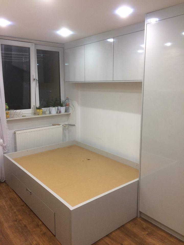 Wnęka z szarą bazą łóżka z szufladami, jasnym drewnianym dnem, białe szafki wiszące i wysoka szafa w zabudowie, widok na okno z roślinami.