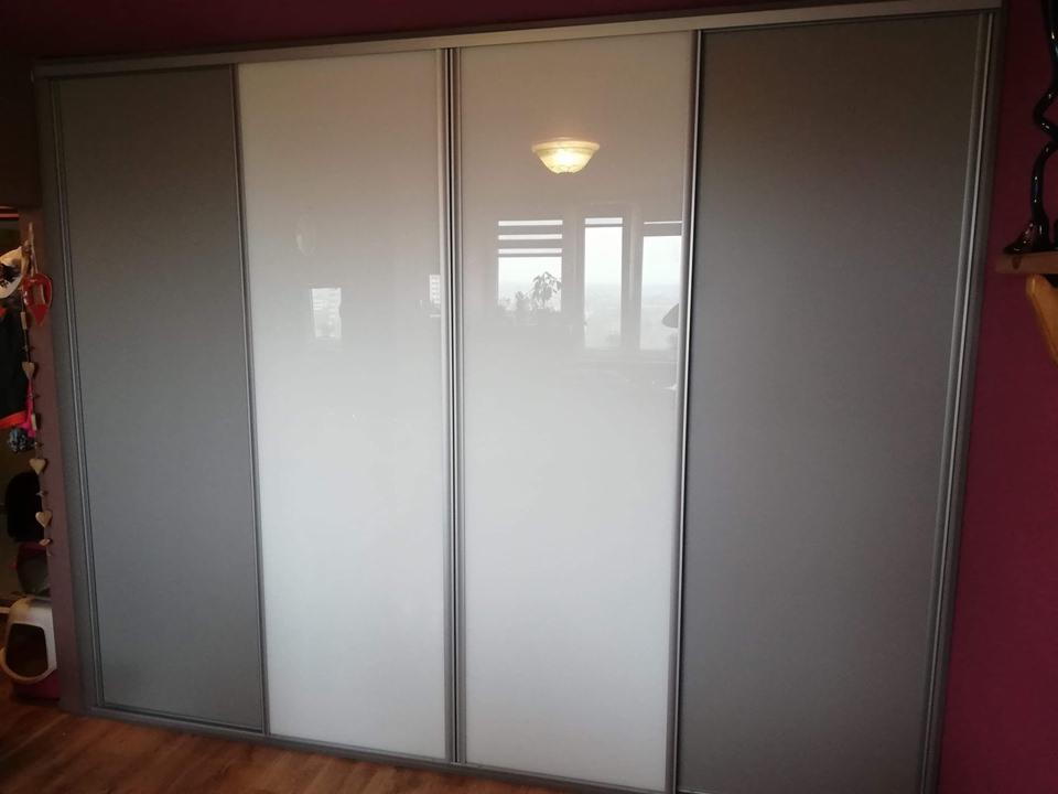 Duża, czterodrzwiowa szafa przesuwna z szarymi i białymi panelami, widoczna na tle drewnianej podłogi i bordowej ściany, z odbiciem okna w białych panelach.