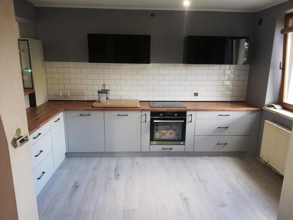 Jasnoszara zabudowa kuchenna z drewnianym blatem i białymi płytkami, widok z perspektywy wejścia do pomieszczenia.