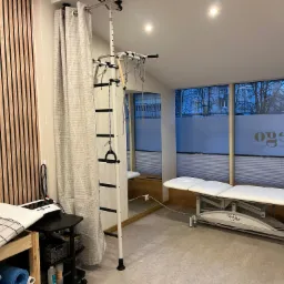 Jasne wnętrze gabinetu z kozetką do zabiegów, drabinką rehabilitacyjną i aparaturą do fizykoterapii, widok z perspektywy pacjenta.