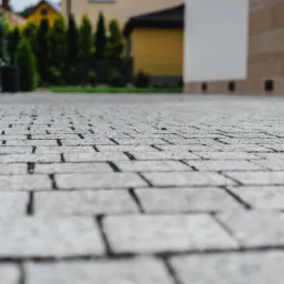 Nowo ułożona szara kostka brukowa tworząca ścieżkę wzdłuż domu z widoczną elewacją i ogrodzeniem w tle, sfotografowana z niskiej perspektywy.