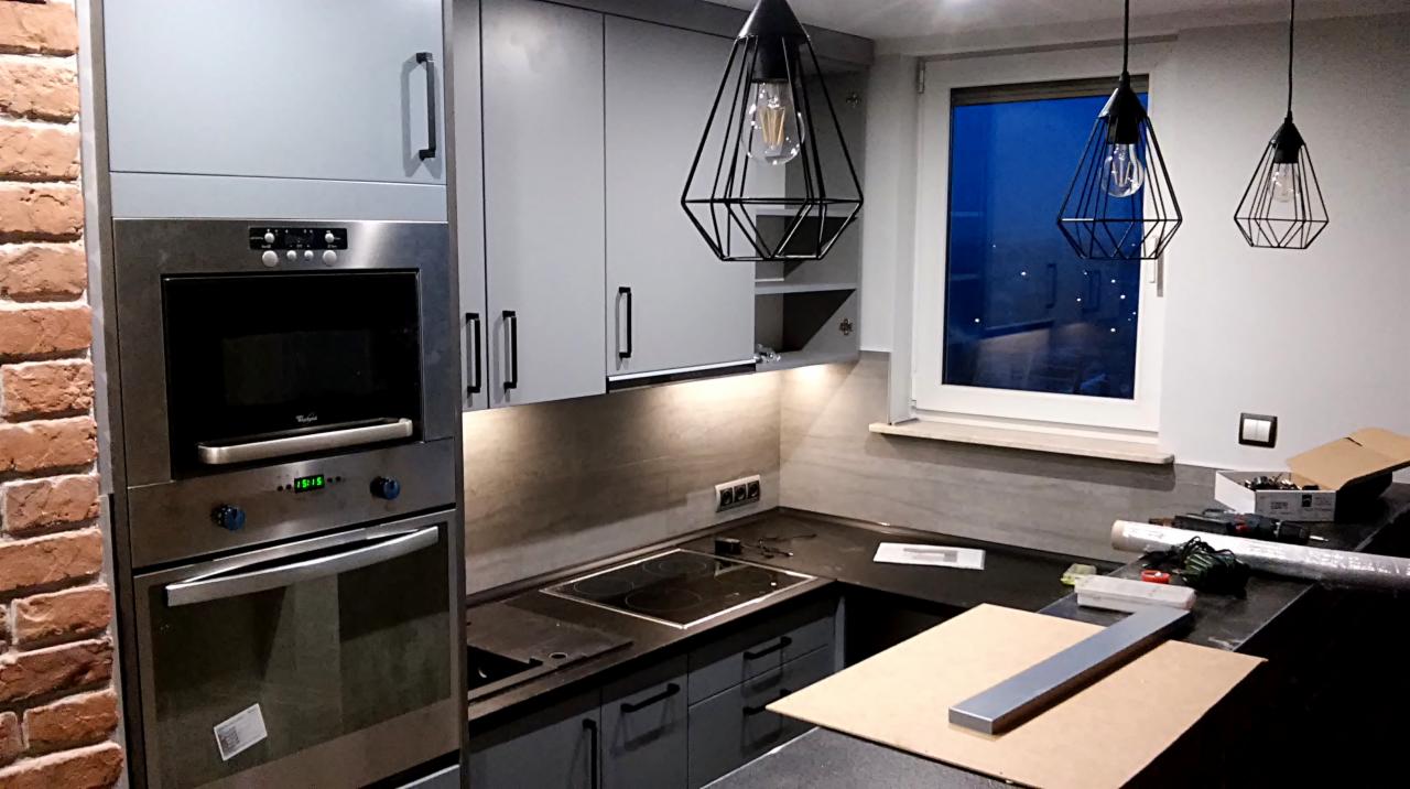 Szara kuchnia z zabudową pod sufit, piekarnikiem i mikrofalówką w zabudowie, czarnym blatem, ceglaną ścianą i trzema lampami w stylu industrialnym, w trakcie montażu.