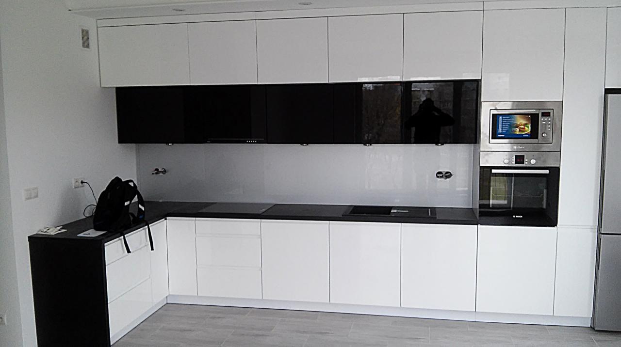 Nowoczesna kuchnia z białymi, lakierowanymi frontami szafek dolnych i górnych, kontrastującymi czarnymi szafkami wiszącymi, czarnym blatem i szarą szklaną ścianą nad blatem, z wbudowanym...