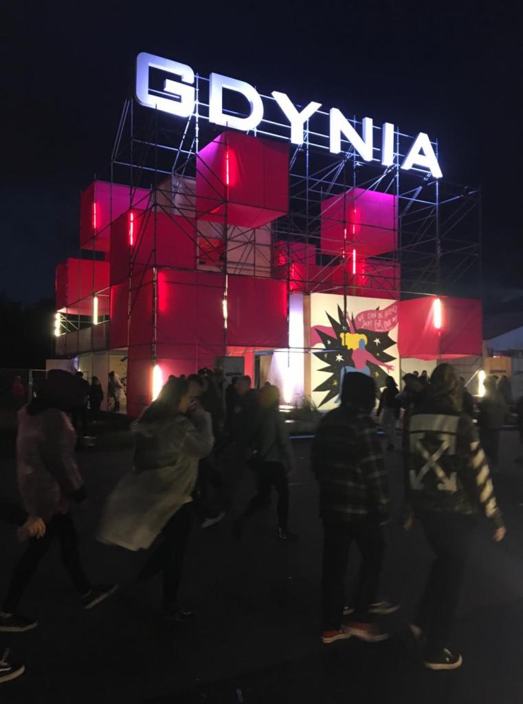 Nocne ujęcie tłumu ludzi przed konstrukcją z napisem 'GDYNIA' i czerwonymi, oświetlonymi kubikami, z muralem w tle.