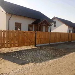 Nowoczesne ogrodzenie drewniane z metalowymi słupkami, front posesji z domami w tle.