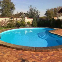 Owalny basen z niebieską wodą i drewnianym obrzeżem, otoczony czerwoną kostką brukową ułożoną w jodełkę, z metalową drabinką i schodkami w wodzie, na tle ogrodu z murem z piaskowca.