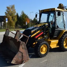 Żółta koparko-ładowarka CAT na kostce brukowej, widok z przodu, z łyżką skierowaną w stronę obiektywu, w tle budynek i drzewa.