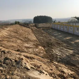 Szeroki wykop w ziemi z widocznymi śladami opon, przygotowany pod budowę, z fragmentem wznoszonej ściany z bloczków betonowych i szklarnią w tle.