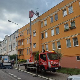 RS INSTALACJE Rafał Stankowiak - Czerwony podnośnik koszowy ustawiony przy pomarańczowym budynku wielorodzinnym, ekipa budowlana przygotowuje materiały na chodniku, prawdopodobnie do prac na dachu.