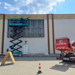 RS INSTALACJE Rafał Stankowiak - Montaż konstrukcji reklamowej na elewacji budynku z użyciem podnośnika nożycowego, widoczny samochód z logo firmy RS-Instalacje i zabezpieczenia terenu.