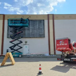 Montaż konstrukcji reklamowej na elewacji budynku z użyciem podnośnika nożycowego, widoczny samochód z logo firmy RS-Instalacje i zabezpieczenia terenu.