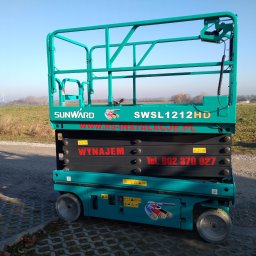 RS INSTALACJE Rafał Stankowiak - Turkusowa podnośnik nożycowy Sunward SWSL1212HD z napisem 'Wynajem' i numerem telefonu, stojący na brukowanej powierzchni, z polami i wiatrakiem w tle.