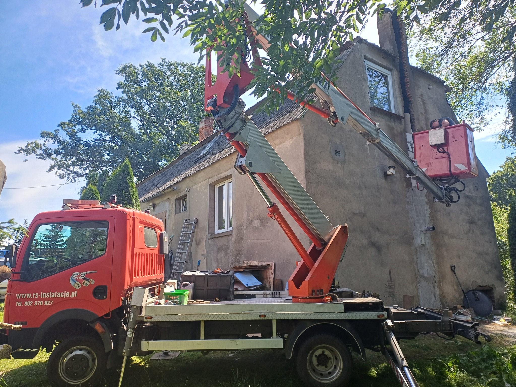 Czerwony podnośnik koszowy na tle starego domu podczas prac na wysokości, pracownik w koszu przy kominie.