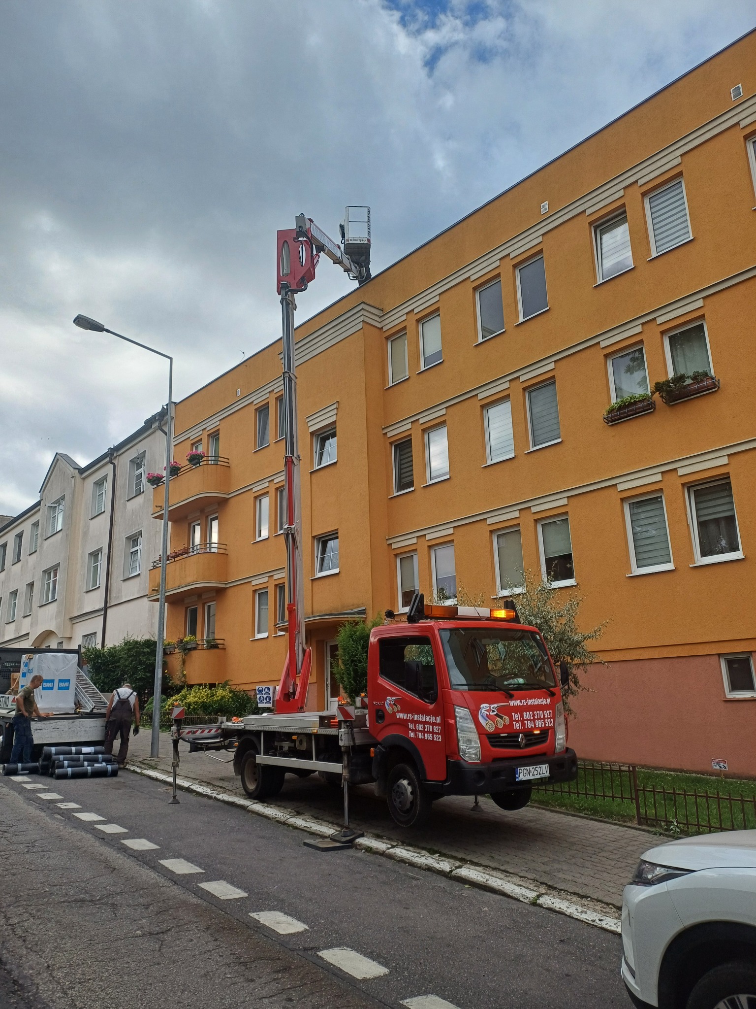 Czerwony podnośnik koszowy ustawiony przy pomarańczowym budynku wielorodzinnym, ekipa budowlana przygotowuje materiały na chodniku, prawdopodobnie do prac na dachu.