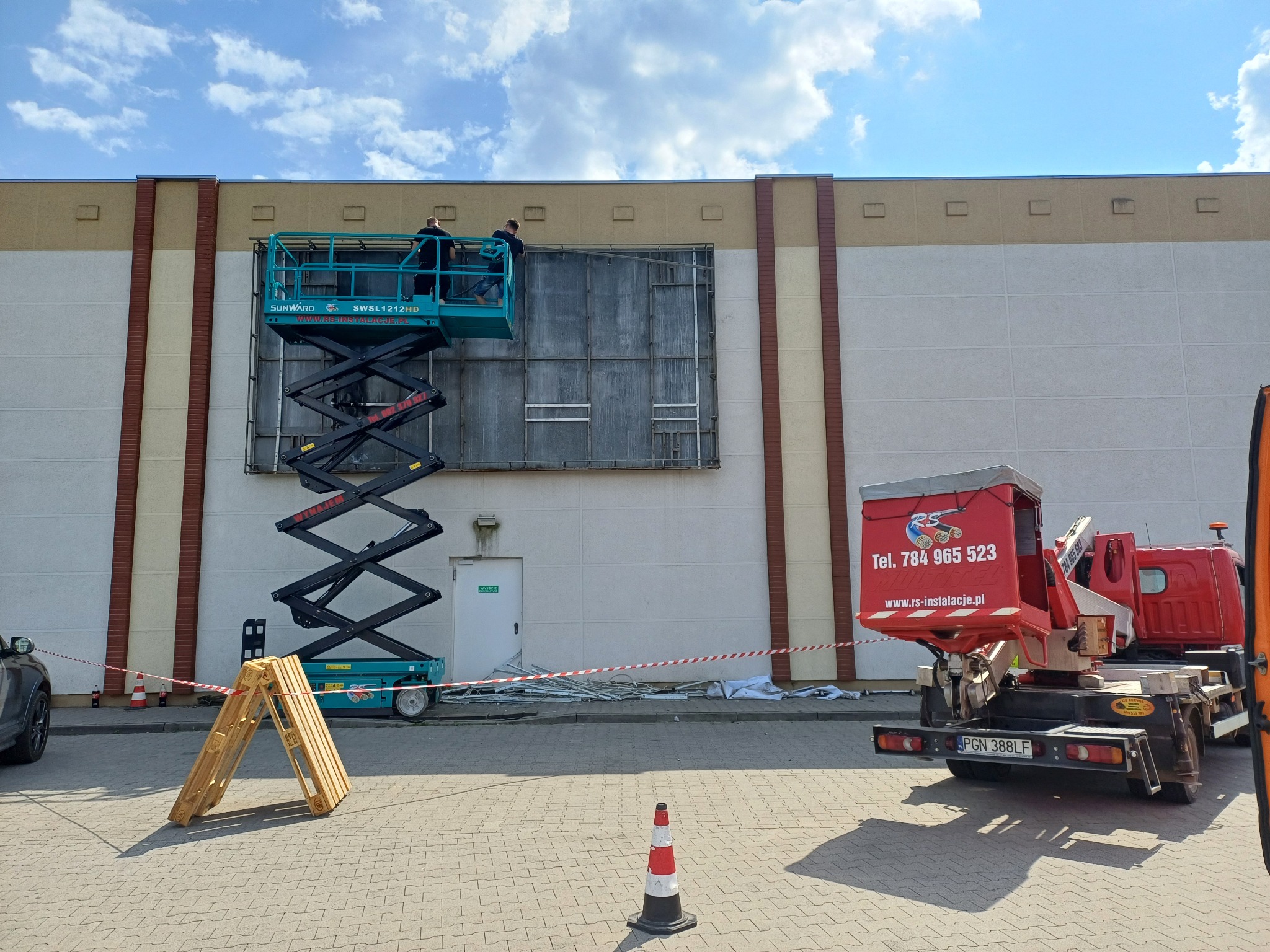 Montaż konstrukcji reklamowej na elewacji budynku z użyciem podnośnika nożycowego, widoczny samochód z logo firmy RS-Instalacje i zabezpieczenia terenu.