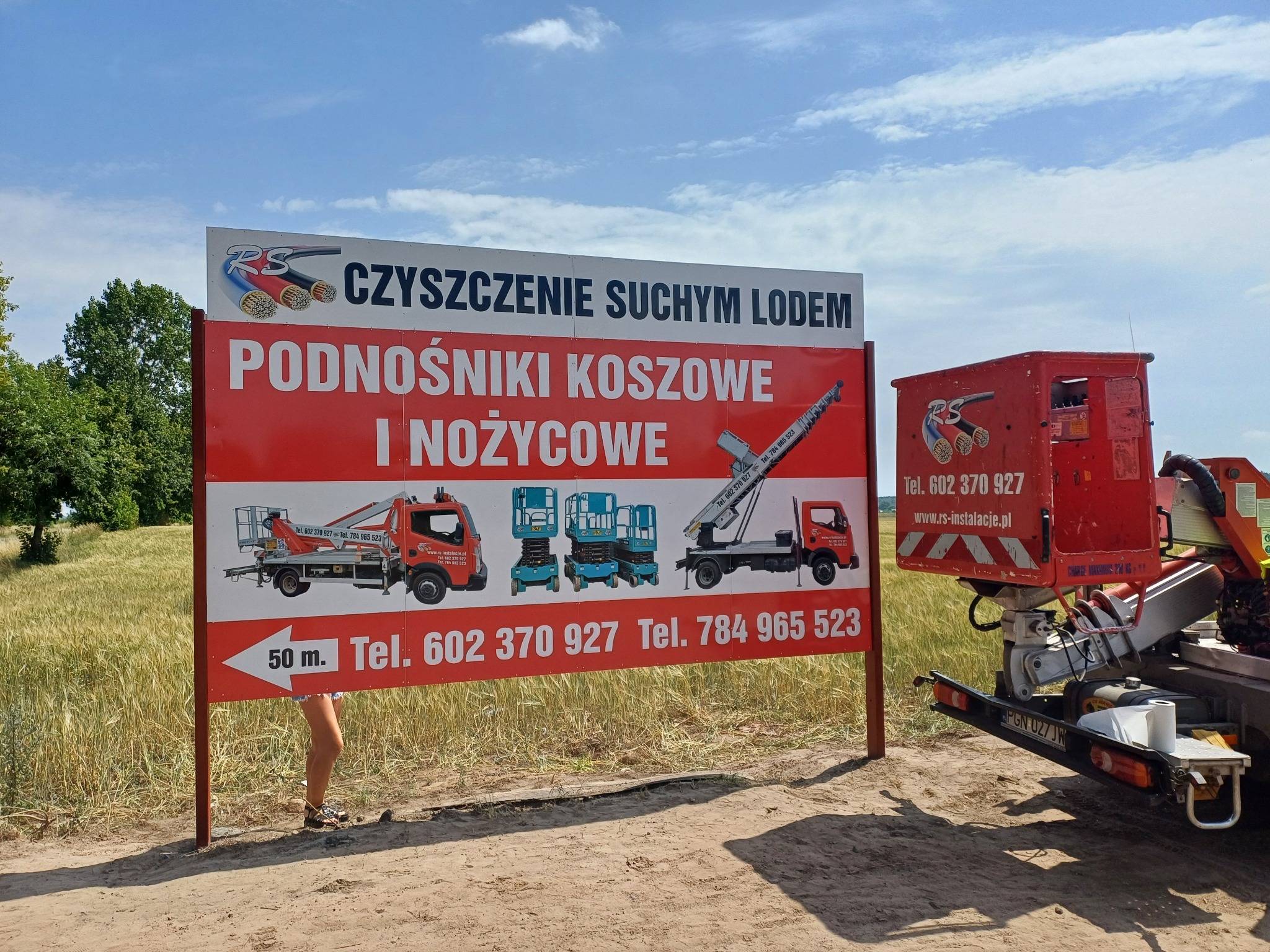 Billboard reklamujący czyszczenie suchym lodem oraz podnośniki koszowe i nożycowe. Na billboardzie widoczne grafiki różnych podnośników. Po prawej stronie zaparkowany podnośnik koszowy z logo firmy.