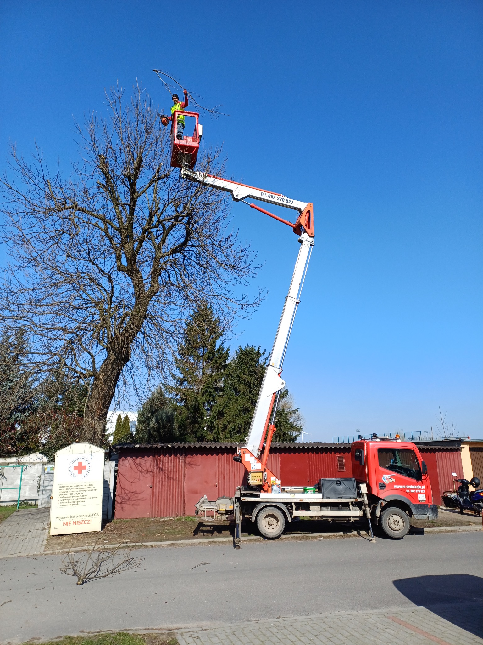 Pracownik w pomarańczowym kombinezonie i żółtej kamizelce na podnośniku koszowym, przycina gałęzie drzewa w słoneczny dzień, na tle błękitnego nieba. Na ziemi leżą odcięte gałęzie, a w tle widoczny...