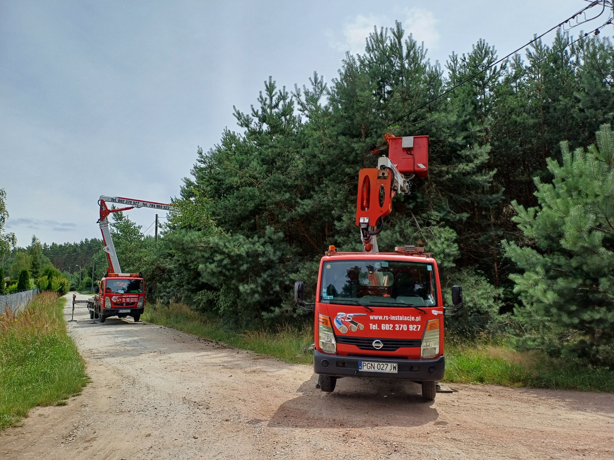 Dwa czerwone samochody z wysięgnikami na polnej drodze, jeden z podnośnikiem w pobliżu linii energetycznych wśród drzew, drugi w oddali.