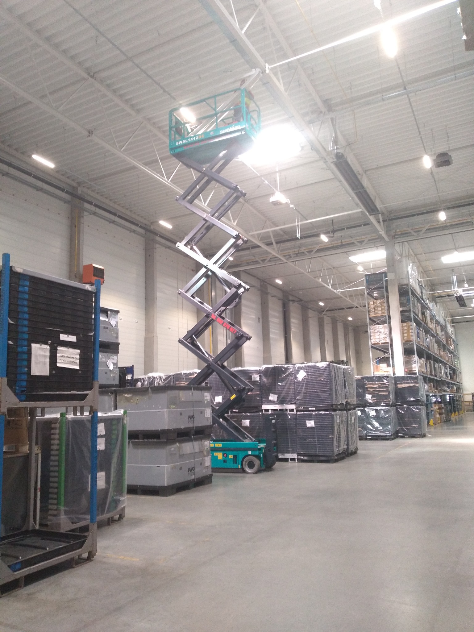 Podnośnik nożycowy w hali magazynowej, widoczne regały z towarem i oświetlenie sufitowe.