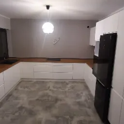 Nowoczesna kuchnia z białymi szafkami i drewnianym blatem, czarna lodówka i okrągła lampa sufitowa, szara podłoga imitująca beton.