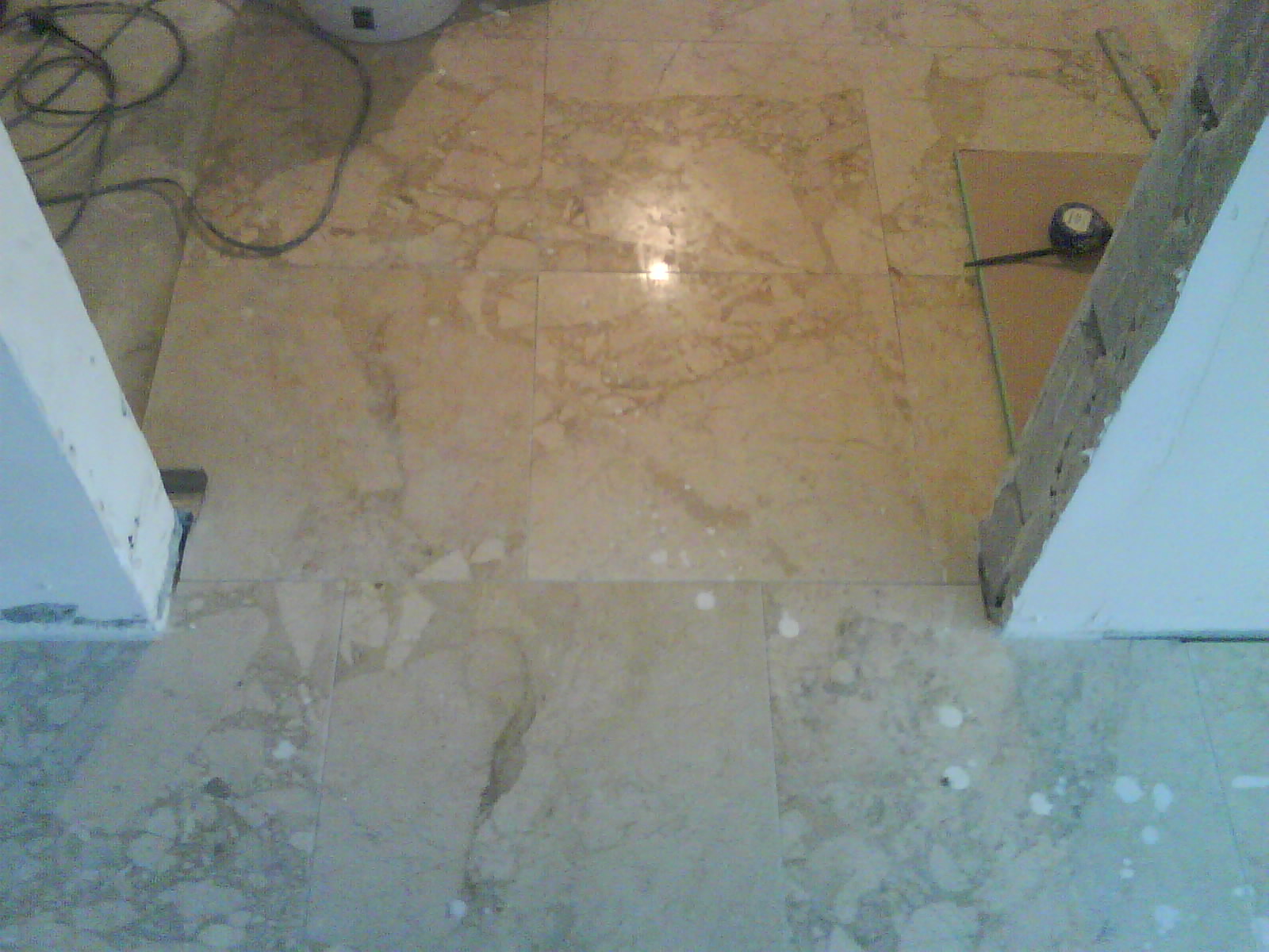 Ułożona marmurowa podłoga w trakcie prac wykończeniowych, widoczne fragmenty ścian, plamy po zaprawie i zwinięta miarka na kartonie ochronnym.