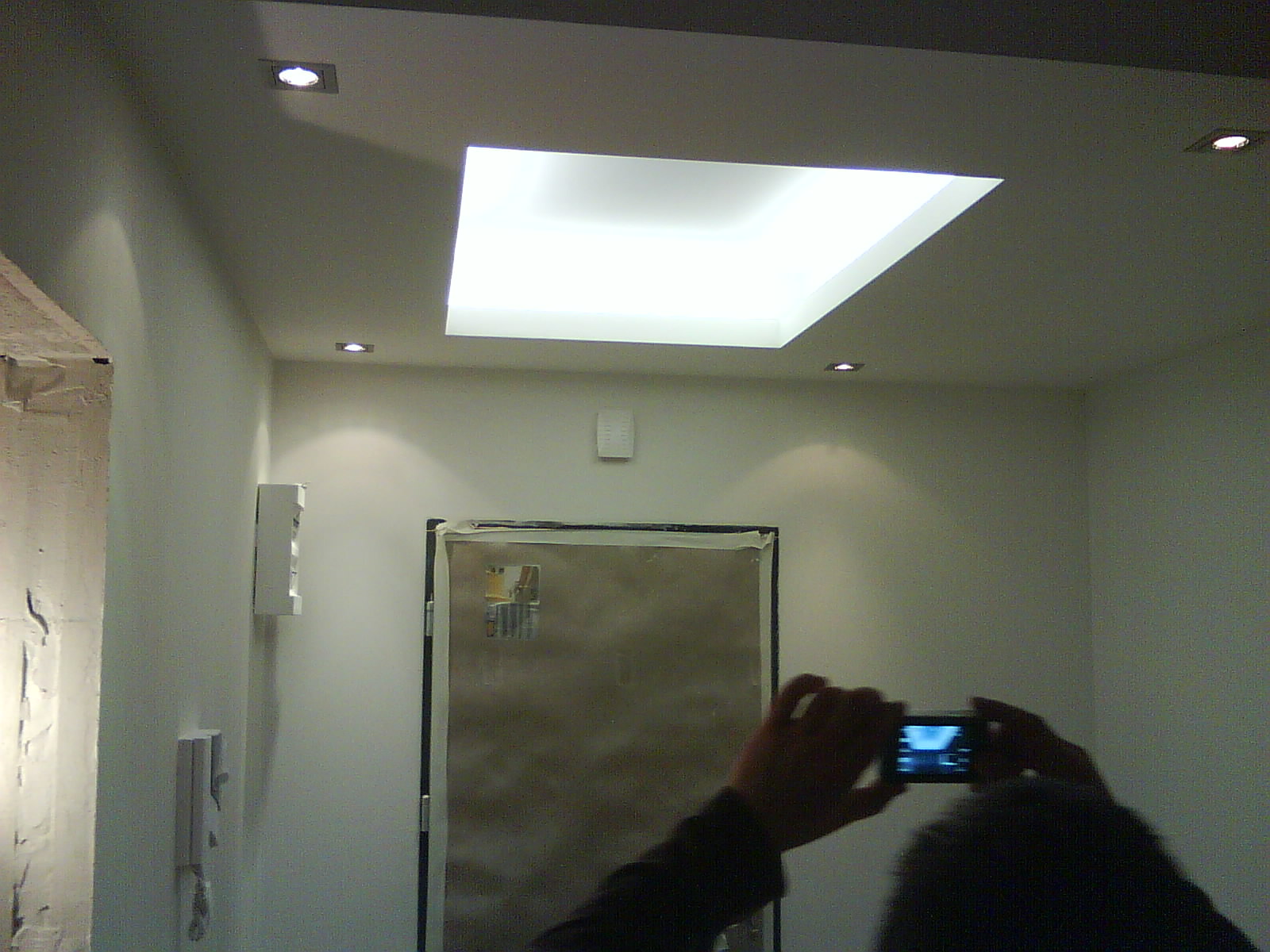 Remontowane wnętrze: zabezpieczone drzwi folią malarską, sufit z oświetleniem punktowym i centralnym panelem świetlnym, osoba robiąca zdjęcie telefonem.
