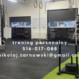 Wnętrze siłowni z wyposażeniem do treningu funkcjonalnego: ergometr wioślarski z napisem #MNTC, liny do wspinaczki, drabinki gimnastyczne, sanie do pchania z obciążeniem, piłki lekarskie na regale...