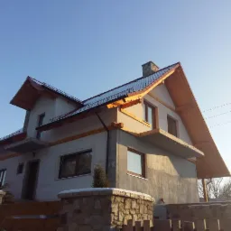 Dwupiętrowy dom w trakcie budowy, z surową elewacją i drewnianymi elementami konstrukcyjnymi dachu, widoczny z niskiej perspektywy w słoneczny dzień.