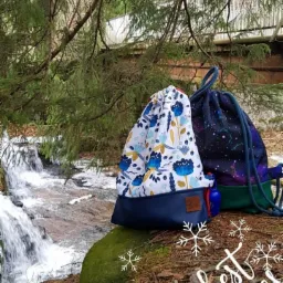 Dwa workoplecaki z tkaniny, jeden z motywem kwiatowym, drugi z motywem galaktyki, stoją na mchu przy strumieniu w lesie, ozdobione grafiką 'best wishes' i śnieżynkami.