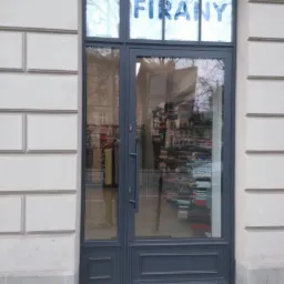 Szare przeszklone drzwi wejściowe do sklepu z firanami, z napisem 'FIRANY' w górnej szybie, widok z zewnątrz na stertę kolorowych materiałów wewnątrz.