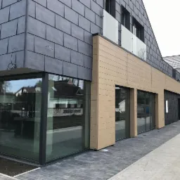 Nowoczesny budynek z elewacją z szarej łupki i beżowych paneli, duże aluminiowe okna narożne z czarnymi ramami odbijające otoczenie, szara kostka brukowa na chodniku, balkon z szklaną balustradą...