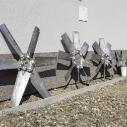 Różnej wielkości metalowe wiatraki z sześcioma łopatami, ustawione w rzędzie przy szarej ścianie z drobnym kamieniem na ziemi, najmniejszy z nich częściowo zasłonięty białym wiadrem.