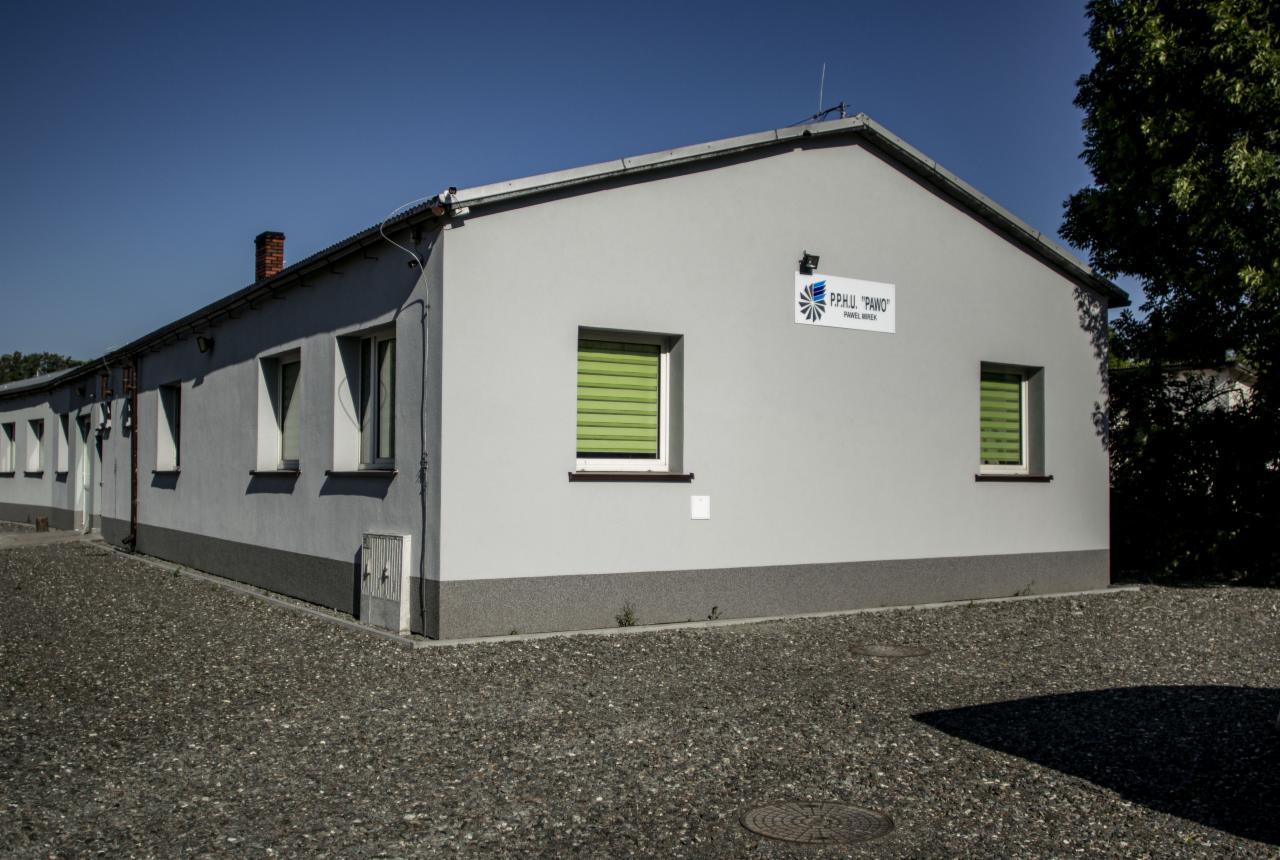 Szary budynek z zielonymi żaluzjami i logo firmy P.P.H.U. 'PAWO' na elewacji, widok z zewnątrz na żwirowy plac.
