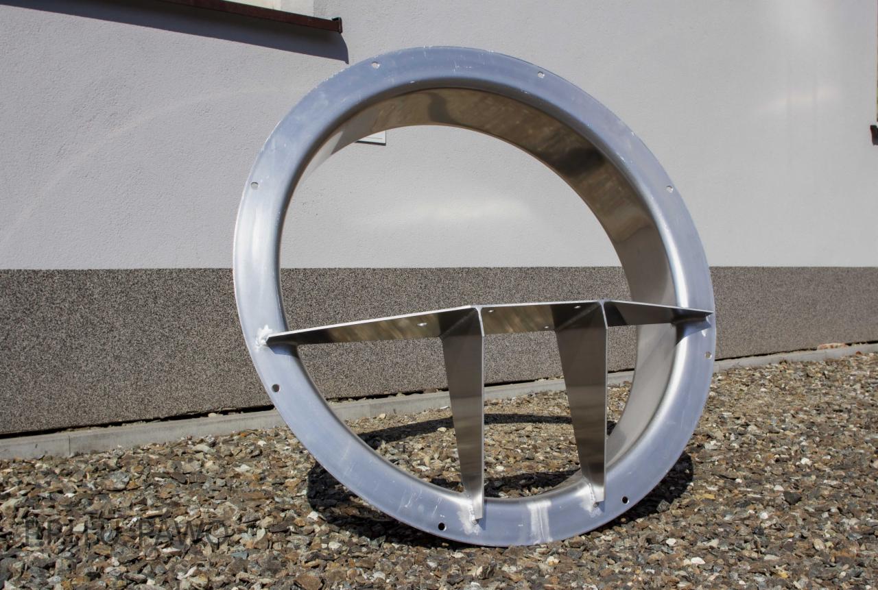 Metalowy pierścień z trzema wspornikami, leżący na żwirze, widoczne otwory montażowe na obwodzie pierścienia.