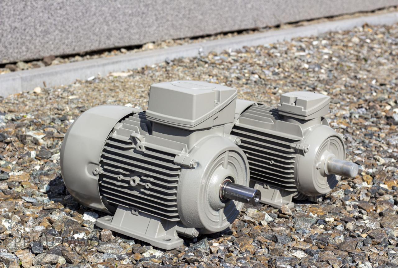 Dwa szare silniki elektryczne na żwirowym podłożu, widoczne żebrowane chłodzenie i wały, betonowa ściana w tle.