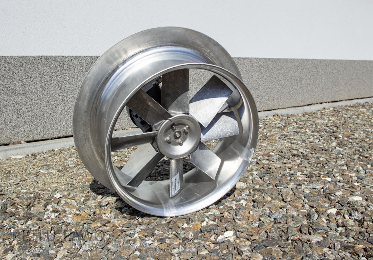 Metalowy wentylator przemysłowy z pięcioma łopatkami i okrągłą obudową, ustawiony na żwirowym podłożu przy szarej ścianie z widocznym cieniem.