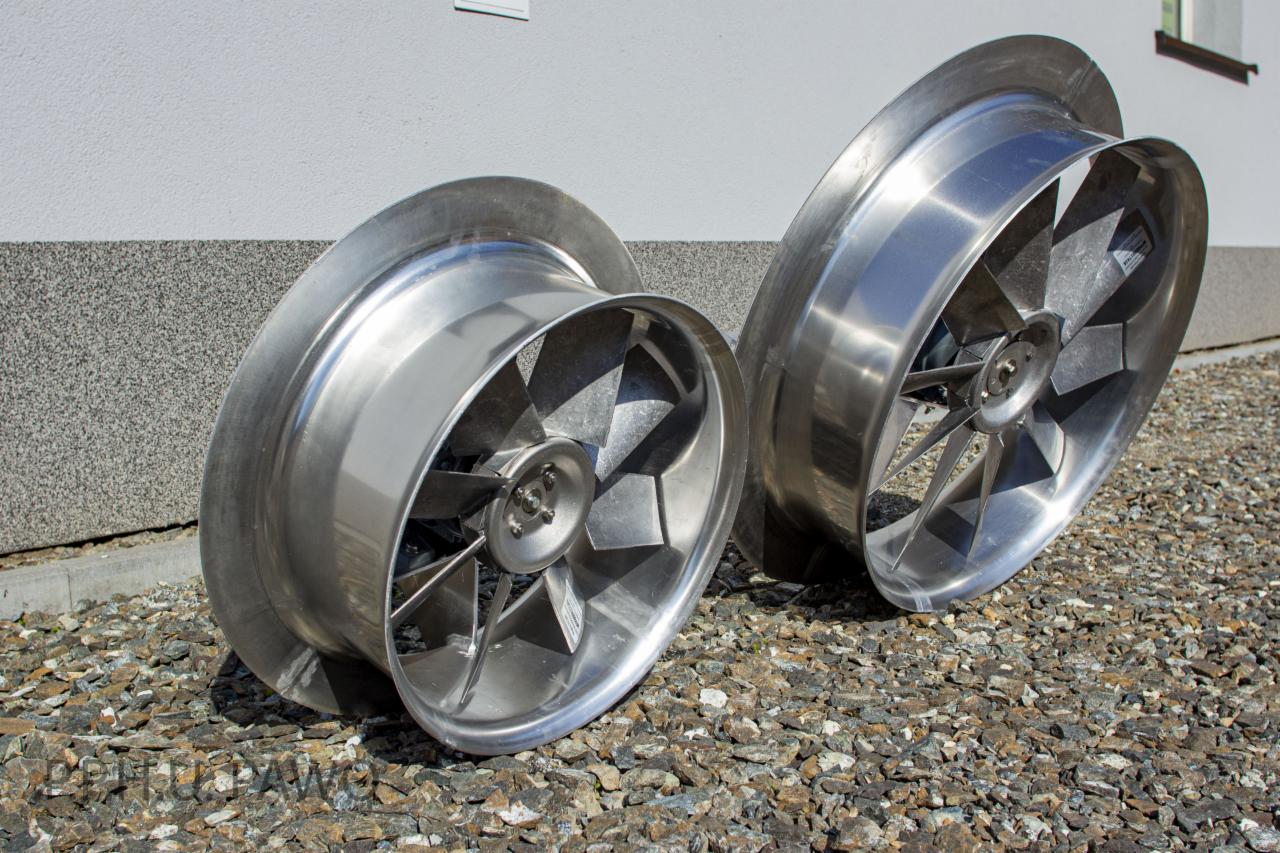 Dwa metalowe wentylatory przemysłowe leżące na żwirze przed budynkiem, widoczne ślady obróbki metalu.