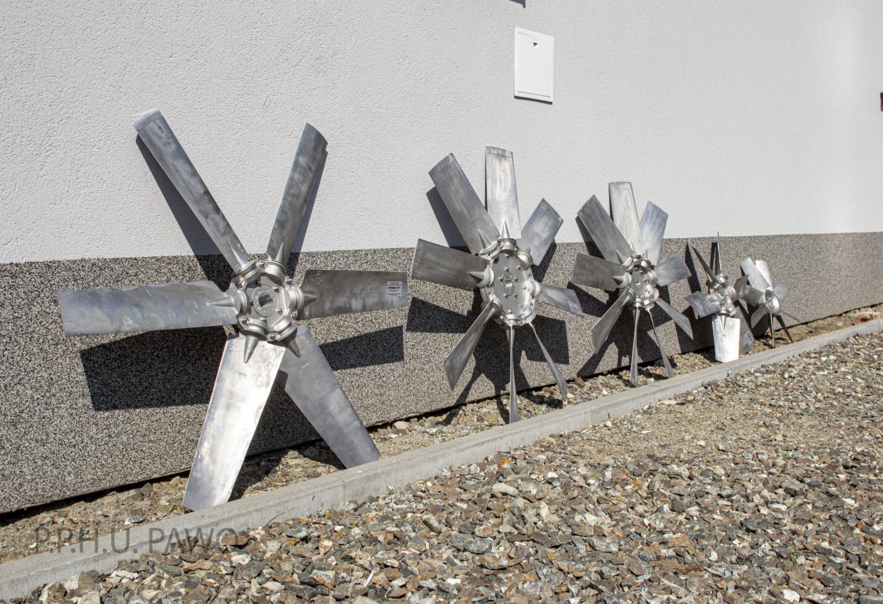 Różnej wielkości metalowe wiatraki z sześcioma łopatami, ustawione w rzędzie przy szarej ścianie z drobnym kamieniem na ziemi, najmniejszy z nich częściowo zasłonięty białym wiadrem.