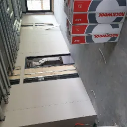 Wnętrze w trakcie wykańczania, widoczne rury wentylacyjne, konstrukcja metalowa, częściowo zamontowane białe płyty, okna oraz paczki materiałów izolacyjnych ROCKWOOL ALU.