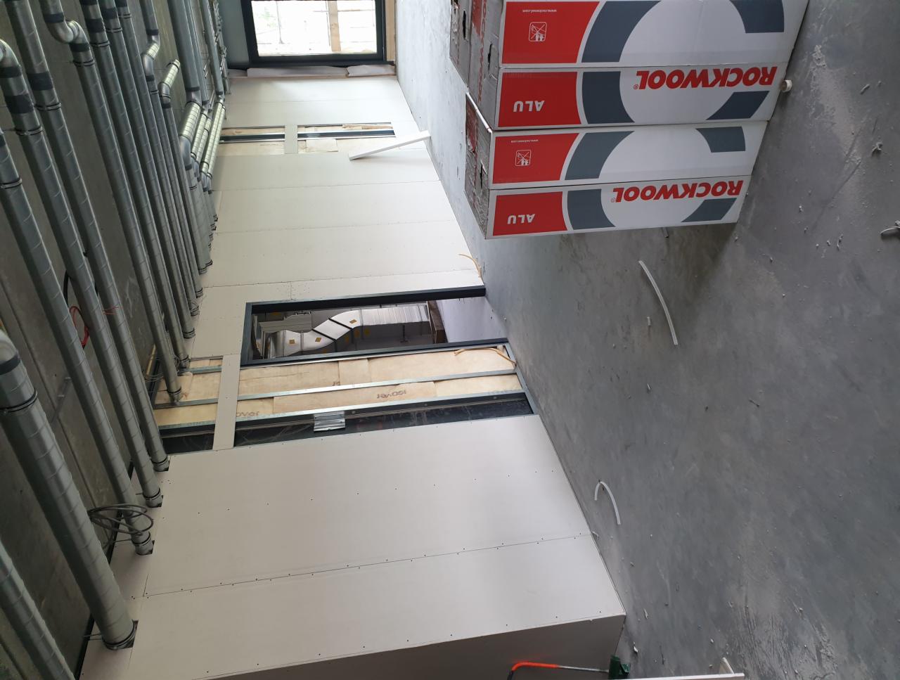 Wnętrze w trakcie wykańczania, widoczne rury wentylacyjne, konstrukcja metalowa, częściowo zamontowane białe płyty, okna oraz paczki materiałów izolacyjnych ROCKWOOL ALU.